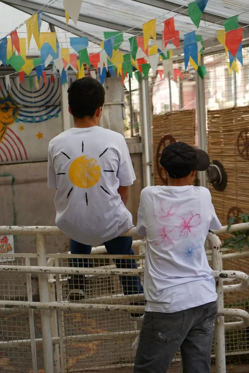 Dois meninos de costas vestindo camisetas brancas pintadas com um sol e flores